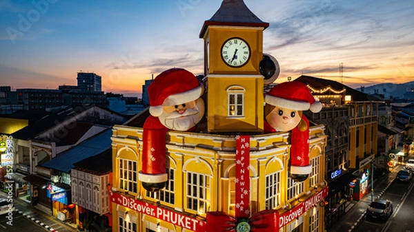 Obraz Phuket,Thailand-December,30,2024: A vibrant scene featuring a historic yellow clock tower adorned with giant Santa Claus and Mrs. Claus decorations during a dramatic evening sky. Happy New Year 2025