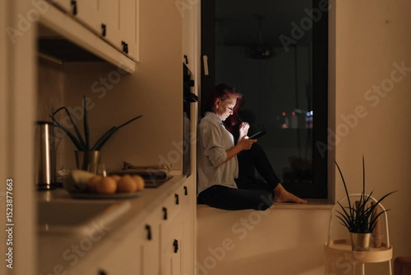 Fototapeta Woman sits in windowlight alone at night looking at glowing smartphone representing blue light at night digital intimacy and emotional solitude during evening scrolling routine in modern lifestyle