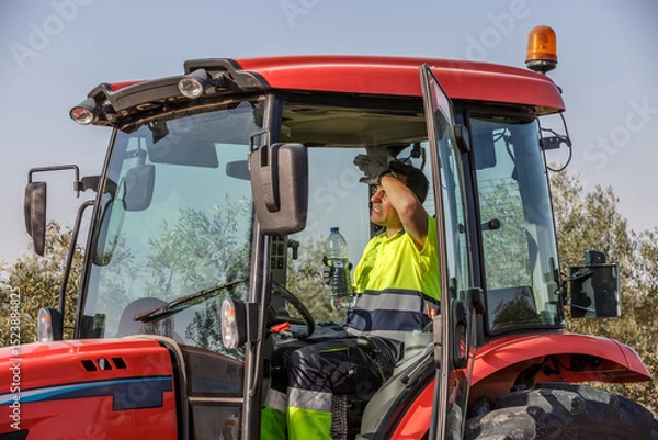 Fototapeta Farm worker in tractor struggles with summer heat while providing safety and hydration