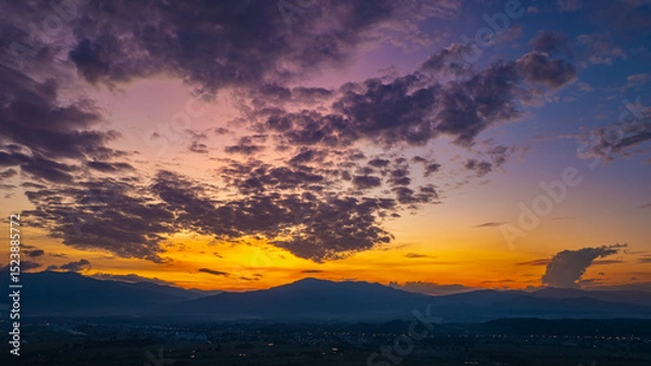 Obraz A breathtaking aerial panoramic view of a vibrant sunset illuminating the sky above a lush green hilltop with a small structure, surrounded by scattered clouds and distant landscapes.