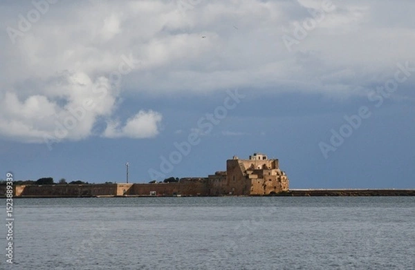 Fototapeta Sunlight falls on ruined stone fortifications on a harbour breakwater. The sky is overcast and the water is grey. The fort and defensive wall are of sandstone.
