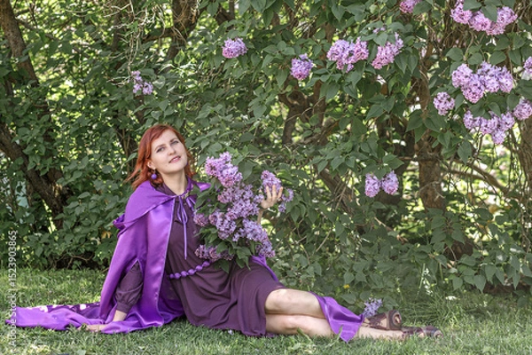 Fototapeta A beautiful young woman with red hair, against a background of blooming lilacs, in a cloak and hood. Medieval fantasy costume. Fairy tale heroine, princess, duchess.