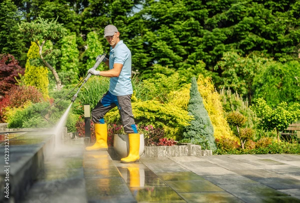 Fototapeta Man Using Pressure Washer to Clean Patio in a Vibrant Garden on a Sunny Day