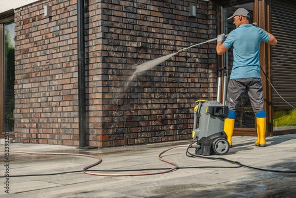 Fototapeta Man Using Pressure Washer to Clean Brick Wall of Modern House During Daytime