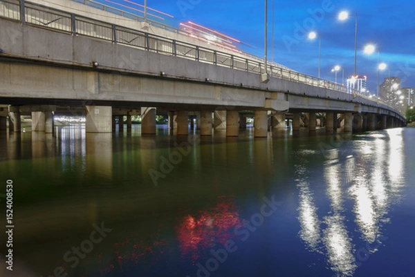 Fototapeta Bridge over water in Linh Dam, Hanoi at dusk showcasing reflections of lights and serene surroundings