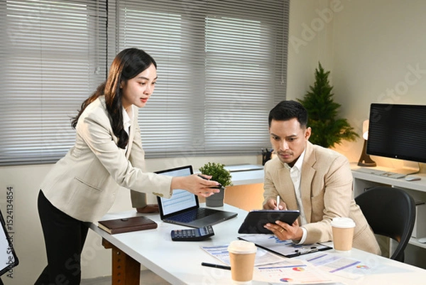 Obraz A woman and a man are sitting at a desk with a laptop and a tablet. The woman is handing the tablet to the man, possibly showing him something on the screen