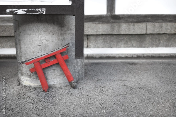 Fototapeta A small, broken red torii gate leans against a concrete pillar on a gray asphalt surface. 