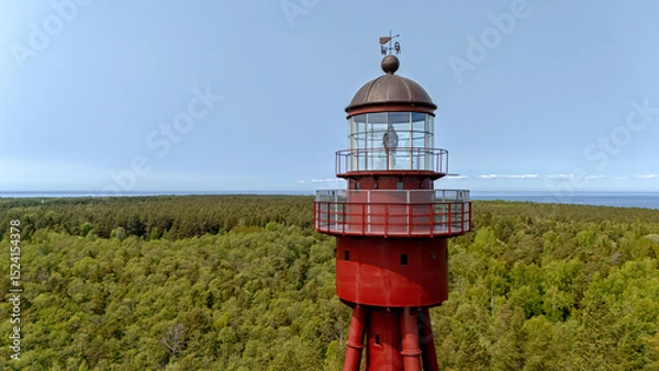 Fototapeta Red lighthouse stands tall above green forest on Ruhnu Island, Estonia during bright daylight