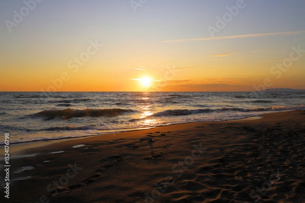 Fototapeta Sandy beach with gentle waves, footprints marking the shore. The sun sets on the horizon, casting a warm orange and yellow hue across the sky, reflecting on the water's surface.