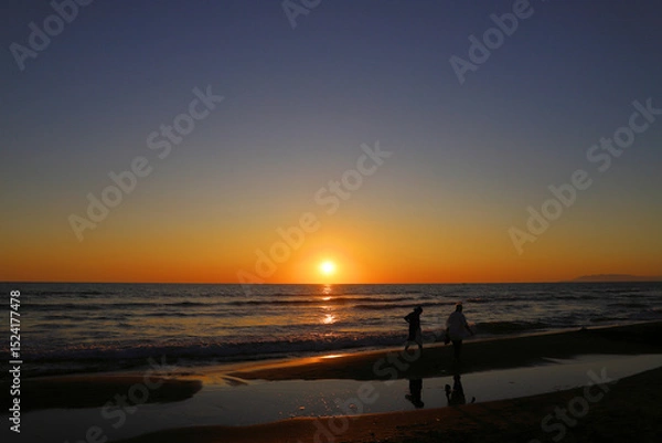 Fototapeta Silhouettes of two people walk along a beach, where gentle waves lap the shore. The sky transitions from deep blue at the top to warm orange near the horizon, as the sun descends.