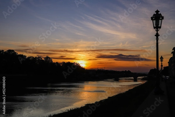Fototapeta Sunset over the Arno River in Pisa, Italy, with an orange and purple sky reflecting on the water. Silhouetted trees line the riverbank on the left, while a walkway with street lamps is visible.