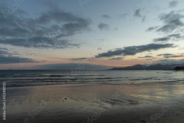 Fototapeta Calm beach scene at dusk with gentle waves lapping against the shore. The wet sand reflects the orange and purple hues of the fading sunset. Wispy clouds are scattered across the sky.