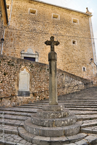 Fototapeta Stone stairs lead to a rustic building constructed with rough, beige stone and small square windows. At the forefront, a stone Celtic cross is mounted on a cylindrical pedestal.