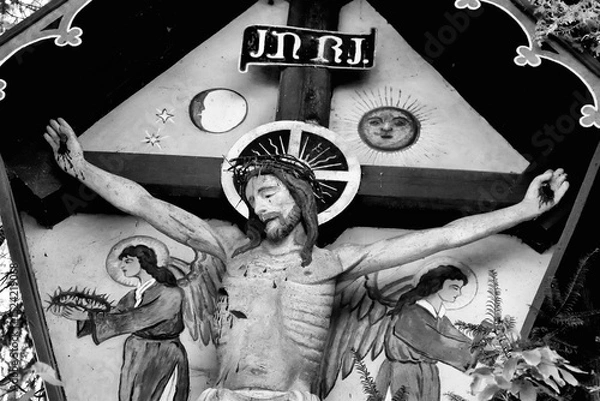 Fototapeta Wooden crucifixion sculpture depicting Jesus Christ on a cross, with a crown of thorns and a serene expression. The plaque above reads "INRI." Flanked by artistic depictions of angels with wings.