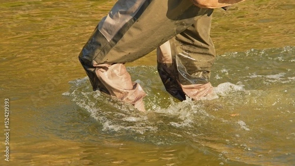 Fototapeta Unrecognizable Fisherman or Angler walks in the wader and wading boots at the slippery rocky rapid river or creek for the salmon and trout spot to fish. Fishing game and sport for fisherman.
