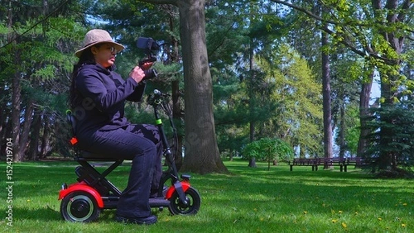 Fototapeta A female videographer person with physical disabilityusing and riding a three wheel wheelchair or e scooter for video recording and content creating. Freedom and independence concept.