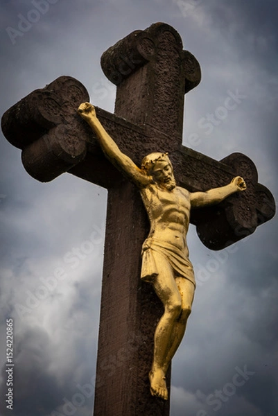 Fototapeta A golden crucifix depicts a figure with outstretched arms nailed to a large, ornate stone cross. The cross is characterized by rounded ends at each arm, resembling a fleur-de-lis design.