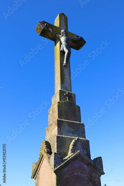 Fototapeta Stone cross featuring a detailed sculpture of Jesus Christ, with weathered textures and moss. Positioned against a clear blue sky, the cross stands atop a robust base.