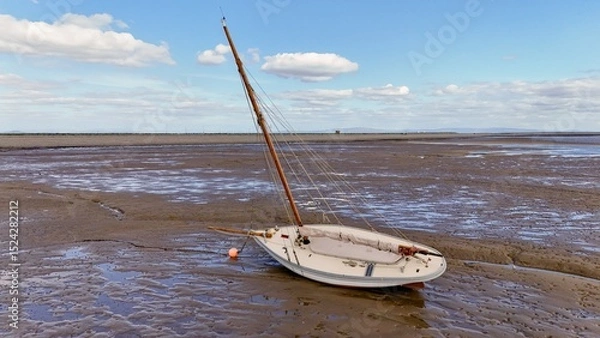 Fototapeta Tilted sailboat resting on muddy flats under calm sky on British coastline.