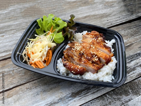 Fototapeta Grilled chicken served on steamed rice, garnished with sesame seeds, accompanied by fresh lettuce, shredded cabbage, and carrot salad with mayonnaise dressing. Presented in a black takeaway box on a r