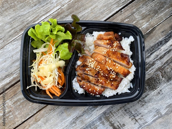 Fototapeta Grilled chicken served on steamed rice, garnished with sesame seeds, accompanied by fresh lettuce, shredded cabbage, and carrot salad with mayonnaise dressing. Presented in a black takeaway box on a r