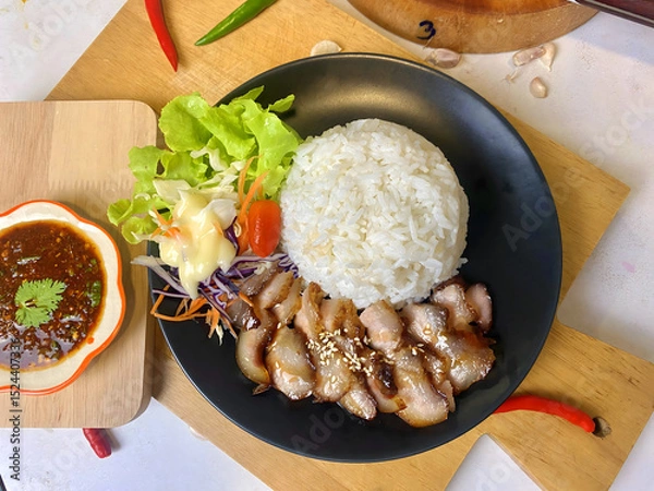 Fototapeta Appetizing Thai grilled pork neck with sesame seeds served with steamed jasmine rice and fresh salad including lettuce, purple cabbage, carrot, and cherry tomato. Presented on a black plate with spicy