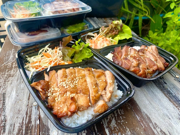 Fototapeta Grilled chicken served on steamed rice, garnished with sesame seeds, accompanied by fresh lettuce, shredded cabbage, and carrot salad with mayonnaise dressing. Presented in a black takeaway box on a r