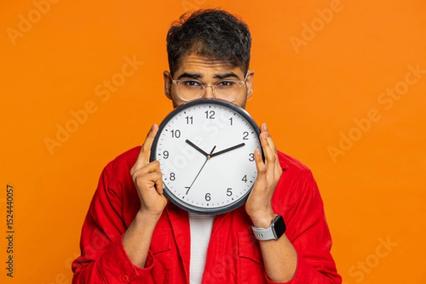Fototapeta Serious strict Indian man holding office clock hiding behind, checking time on watch, running late to work, being in delay, deadline, obscuring face. Arabian guy isolated on orange background indoors