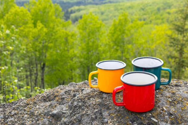 Fototapeta Three metallic tea cups—red, blue, and yellow—on ancient mountain rocks , set against a breathtaking view of a forested valley in sun light. Camping and hiking vibes of adventure.