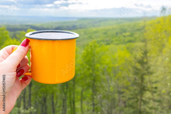Fototapeta A woman's hiker hand holding a bright yellow metallic cup on the top of a mountain in front of breathtaking view of a forested valley in sun light. Camping and hiking vibes of adventure.