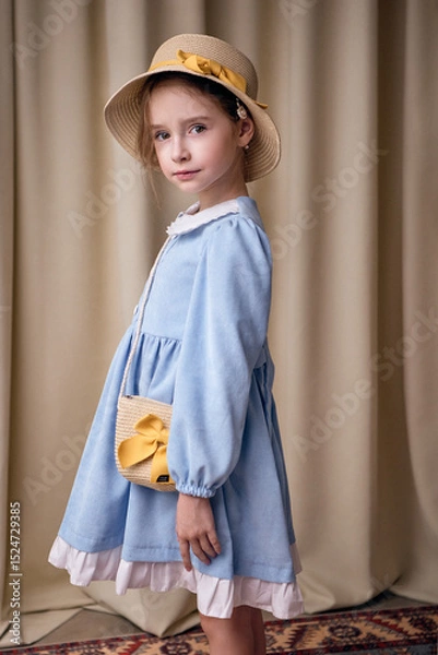 Fototapeta A cute girl with a serious expression poses half-turned. Her hair is tucked under a straw hat. The model is wearing an elegant velvet dress with frills on a beige background