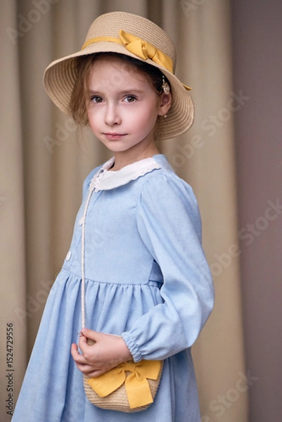 Fototapeta A seven-year-old girl with soft features looks thoughtfully at the camera. She is wearing a set of straw accessories, a woven hat with a yellow ribbon and a shoulder bag. The model in a romantic look 