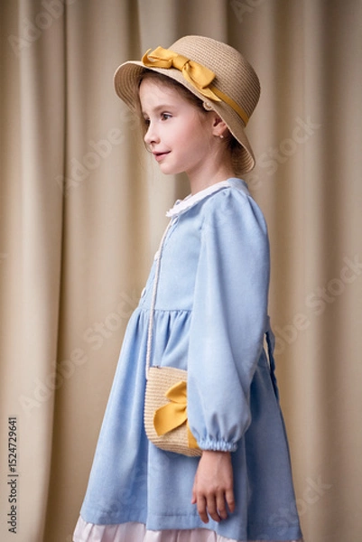 Fototapeta A sweet young girl in a romantic dress stands sideways and looks to the left. On her head she has a straw hat with a yellow ribbon and bow. Shot from the bottom up against a plain light curtain.
