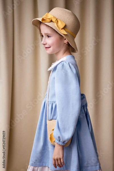 Fototapeta A funny little girl in profile on a fabric background. The child is looking to the left and smiling in a cheerful mood. The model is wearing a dress and a hat with a ribbon.