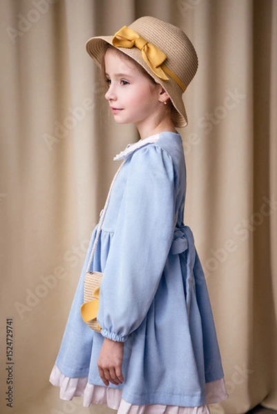 Fototapeta A little girl stands in profile in a light blue dress with white frills at the bottom. On her head she has a straw hat with a yellow ribbon and bow. A light curtain is visible in the background.