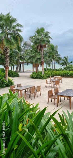 Fototapeta Outdoor Dining Area with Wooden Tables and Chairs Under Palm Trees Near the Sea. Tropical Resort Restaurant Setup.