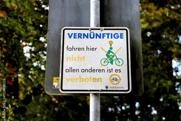 Fototapeta A prohibition sign is mounted on a metal pole, surrounded by lush green foliage. The sign features a diagram of a cyclist with an overlaid red cross, topped by a lightbulb icon.