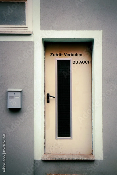Fototapeta Cream-colored door with a vertical window panel, set in a gray wall. Above the door is a German sign: "Zutritt Verboten DU AUCH" meaning "No Entry YOU TOO." A dark handle is on the left.