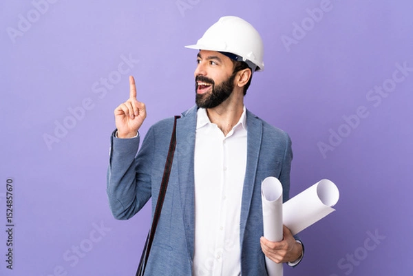 Fototapeta Young architect man with helmet and holding blueprints over isolated purple background thinking an idea pointing the finger up