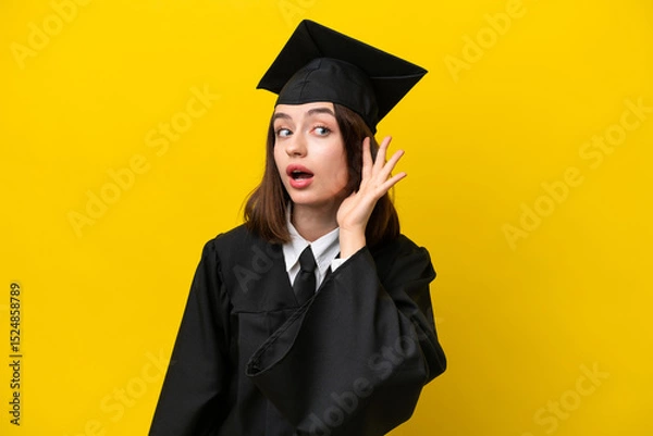 Fototapeta Young university graduate Ukrainian woman isolated on yellow background listening to something by putting hand on the ear