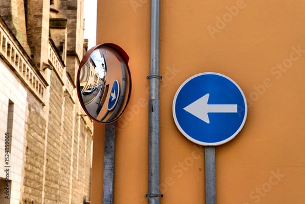 Fototapeta A blue circular traffic sign with a white left-pointing arrow is mounted on an orange wall. Beside it, a convex mirror reflects the street view, assisting drivers with visibility around the corner.
