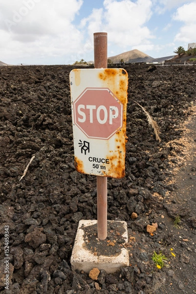 Fototapeta Rusty stop sign on a volcanic landscape, set in black lava rock on Lanzarote, Canary Islands, Spain. The sign indicates a crossing 50 meters ahead ("CRUCE 50 m").