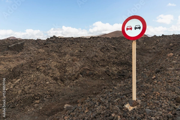 Fototapeta A road sign stands on rugged, dark volcanic soil in Lanzarote, Canary Islands, Spain. The sign features a red circle with symbols of two cars, indicating a potential traffic restriction.