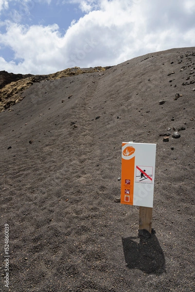 Fototapeta A wooden post supports a warning sign against sliding or rolling, placed on black volcanic sand. The sign features an orange and white color scheme and pictograms indicating prohibited actions.