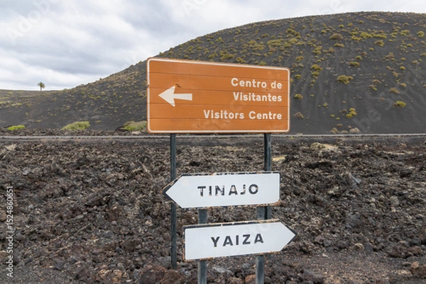 Fototapeta Signpost located on a rugged volcanic landscape in Lanzarote, Canary Islands, Spain. The sign is mounted on a metal pole and consists of three directional panels. The top panel is brown.