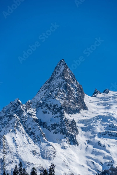 Fototapeta Majestic snow-capped mountain peak stands tall against clear blue sky, showcasing rugged beauty of winter landscape. Karachay-Cherkessia. Dombay 2025. Nature concept for design.