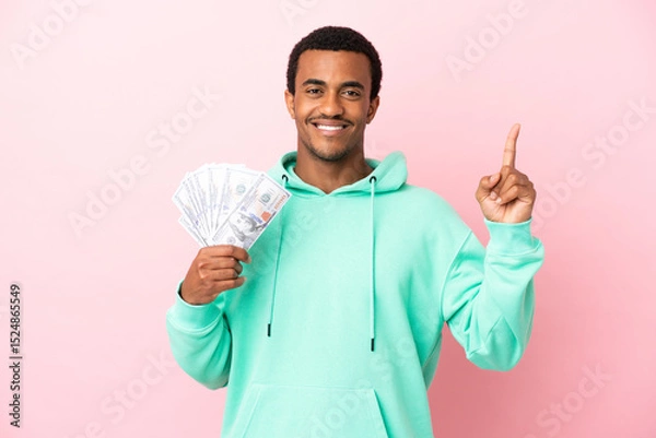 Fototapeta Young man taking a lot of money over isolated pink background pointing up a great idea