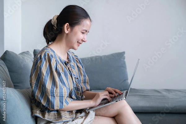 Fototapeta A woman sits on a couch, smiling while using a laptop, embodying a relaxed yet productive atmosphere in a light-filled room.