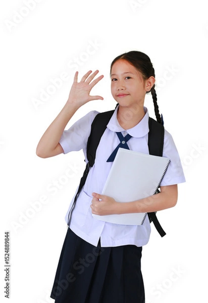 Obraz Smiling Asian student girl in school uniform waving hand hello while carrying backpack bag and holding a book against white background.