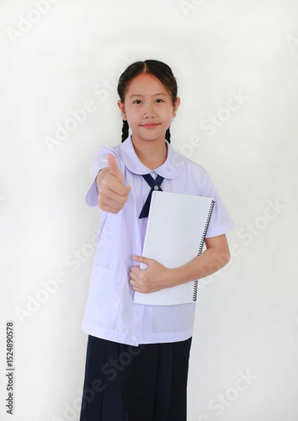Obraz Portrait of Asian student girl in school uniform showing thumb up finger very good gesture while holding a book against white background.